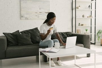 pretty african american girl using laptop and holding credit card while sitting on sofa at home