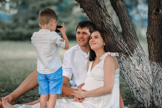 Happy family relaxes in nature under a tree in the afternoon. The child takes pictures of his father and pregnant mother.