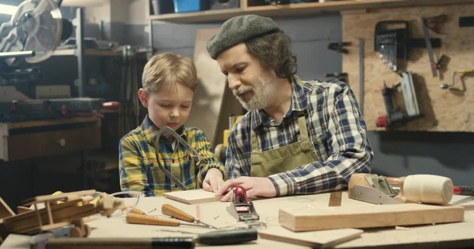Caucasian grandfather carpenter teaching small cute grandson to file with saw in workshop. Old woodman showing little boy how to sawing a timber and work with wood. File using concept.