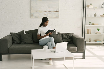 attentive african american freelancer reading notebook while sitting on sofa near table with laptop