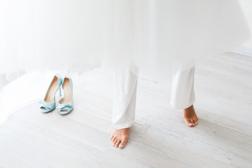 Barefoot bride in white trousers stands next to blue wedding shoes