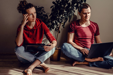 Real modern couple in casual clothes sitting on floor working from home on laptops. Thoughtful woman in red hoody with netbook work on her project, happy man in t-shirt use notebook computer.