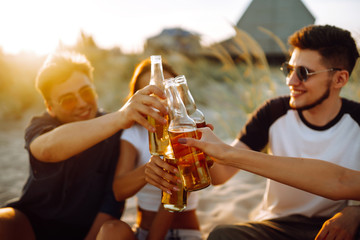 Group of young friends sitting together at the beach talking and drinking beers at sunset. Summer holidays, vacation, relax and lifestyle consept.