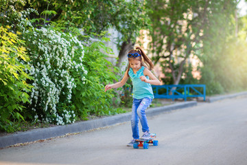 Fototapeta premium A little girl in jeans and sunglasses rides a skateboard around the yard in spring or summer. Children's sports at home