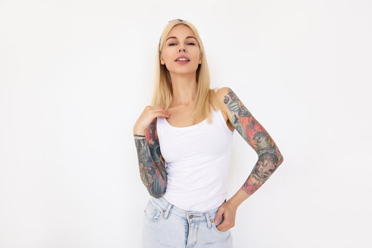 Beautiful young tattooed blonde woman with casual hairstyle raising hand to her hair while looking at camera, isolated over white background in headband and casual wear