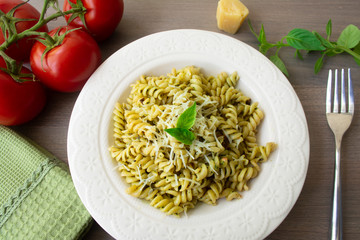 Traditional Italian vegetarian pasta fusilli with fresh homemade basil and pine nut pesto sauce and topped with grated Parmesan cheese. Tomato, basil, parmesan cheese, napkin and fork in background