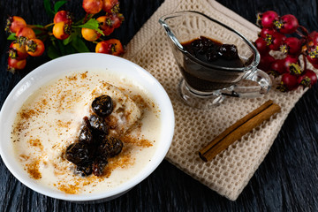 Sour-milk yogurt dessert, with thermostatic cooking, decorated with ripe cherry berries in syrup, on a dark background with jam in sauce and a stick of cinnamon, next to sprigs of fresh red berries