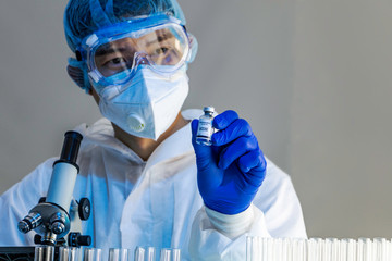 Asian male researcher in full Personal Protective Equipment uniform is holding small bottle of COVID-19 vaccine example. Coronavirus vaccine in scientist's hand under trials is almost ready for human