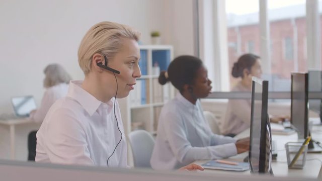 Medium shot of diverse women wearing white shirts and headsets sitting at computers in the office and talking to costumers by telephone