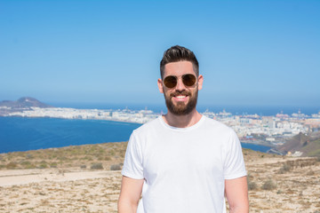 Handsome man with sunglasses, beard, and a white shirt on the top of the mountain from where you can see the edge of the island and the ocean, illuminated by sunlight.