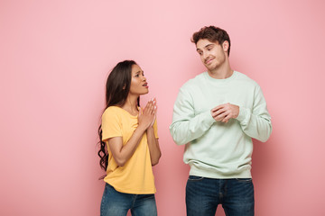 Obraz premium pretty african american girl with praying hands looking at smiling, skeptical boyfriend on pink background