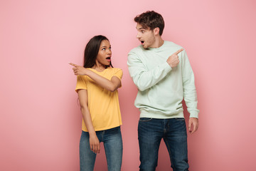 shocked interracial couple looking at each other and pointing away with fingers on pink background
