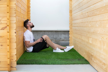 A handsome man sitting on the artificial grass between two wooden cabins.