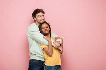 happy guy embracing smiling girlfriend while looking away on pink background