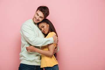 beautiful african american girl looking at camera while happy boyfriend embracing her on pink background