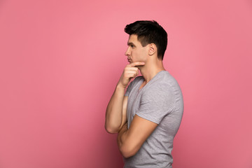 A lot to think about. Close-up photo of an attractive man in a grey t-shirt, who is standing in profile and thinking of something, while touching his chin with a right hand.