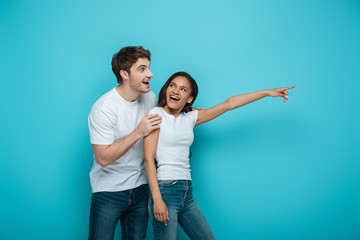 surprised man touching shoulder of smiling african american girl pointing with finger on blue background