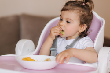 Portrait of happy young baby girl in high chair eating exotic fruits. Healthy nutrition for kids