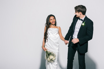 elegant bride groom holding hand of happy african american bride with wedding bouquet on white background