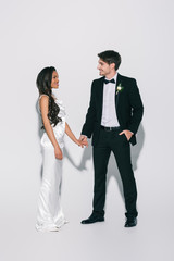 full length view of happy, elegant interracial newlyweds holding hands and looking at each other on white background