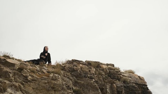 beautiful woman in black dress is sitting on edge of cliff at top of mountain.A young woman enjoys mountain scenery and windy weather.big lake in mountains