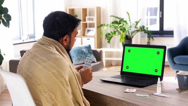 healthcare, technology and people concept - sick indian man in blanket with medicine pills having video call on laptop computer with chroma key green screen at home
