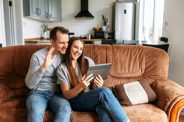 Obraz premium Couple in love are sitting on the couch at home and using a tablet for talking online, video meeting with a friends. They are laughing and waving hello at webcam