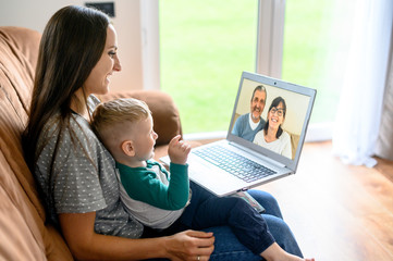 Little grandson is talking via video call on laptop with a grandparents. A young mother with a baby son sit on a sofa at home and have a video meeting grandparent are on a laptop screen