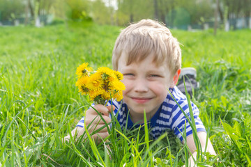 A blond European boy is lying on the grass with a bunch of yellow dandelions.
