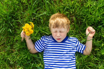A blond European boy is lying on the grass with a bunch of yellow dandelions. The child fell and bumped, the boy is hurt.