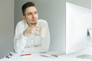 Young man in white shirt with cup working at the desktop with a computer in office. Worker on grey background