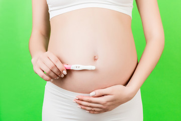 Close up of positive pregnancy test against pregnant woman's belly at green background. Future mother in white underwear. Baby expecting. Copy space