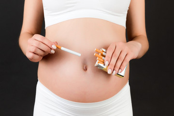 Close of of pregnant woman in white underwear holding a pack of cigarettes at black background. Bad habbit. Toxic influence on a baby