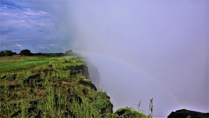 Miracle over Victoria Falls. A rainbow appeared in a thick mist of water spray. On the steep slopes of the gorge, bright green grass. There are clouds in the sky.