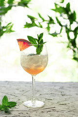Glass of cocktail with grapefruit mint and ice on table against green leaves.