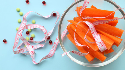 a transparent plate with chopped carrots, a measuring tape, is on a light blue background, next to red and green pills, vitamins, dietary supplements. Weight loss, proper nutrition, vitamins of group 