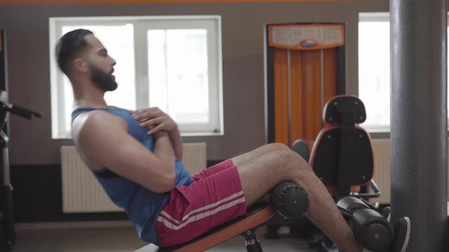 Side view of young Middle Eastern man pumping press using fitness equipment. Workout of strong handsome sportsman in gym. Exercising, sport, healthy lifestyle, strength.