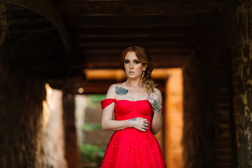 Obraz premium Attractive redhead tattooed woman in red dress and diadema posing on blurred medieval castle background