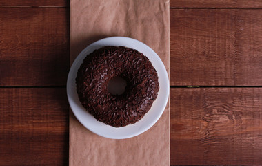 Chocolate donut with white ceramic plate or saucer and craft paper napkin on brown wooden table. Top view. Bakery, dessert, pastry concept