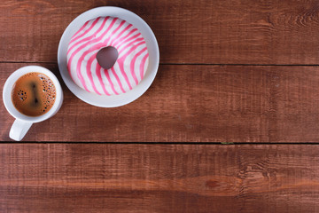 Donut with pink and white glaze and cup of black coffee on brown wooden table. Top view, copy space. Bakery, coffee shop menu, breakfast, break concept
