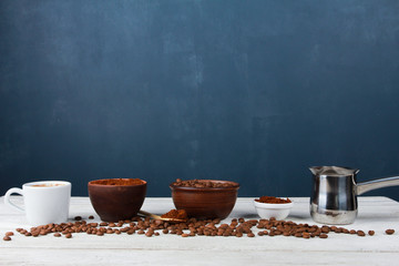 Espresso cup, metal Turkish pot, roasted Arabica beans in clay bowls, coffee powder on white table against dark blue wall. Side view, copy space. Coffee shop, morning, baristas workplace concept