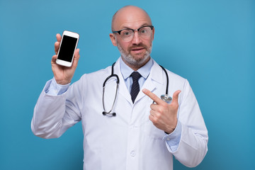 Mature male doctor shows blank screen of smartphone. Studio shot on blue wall.