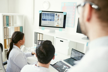 Rear view of doctors looking at computer monitor and talking to their colleague during online...