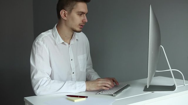 Young man in white shirt working at the desktop with a computer in office. Worker on grey background