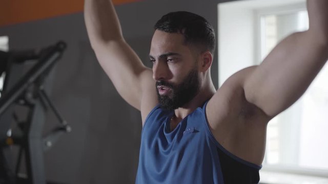 Close-up of confident perspiring sportsman training shoulder and back muscles in gym. Serious handsome Middle Eastern man using pull down machine for muscular built. Sport, health, lifestyle.