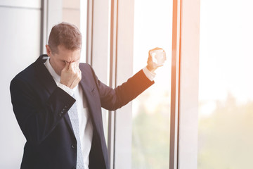 Portrait of serious mature businessman emotional stress working at office hand holding coffee cup. sad standing by the window. Headache migraine. 
