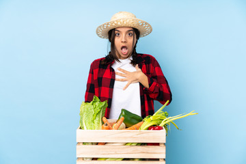 Young farmer Woman holding fresh vegetables in a wooden basket surprised and shocked while looking...