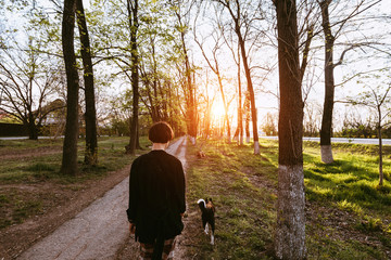 Young woman with a dog walks among the trees along the path at sunset