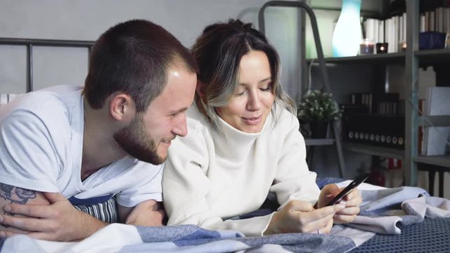 Happy Couple Using Smartphones In Bed At Night. Young attractive Caucasian just married man and woman sitting in their bed late at night and playing on their smartphones while taping, then looking at 