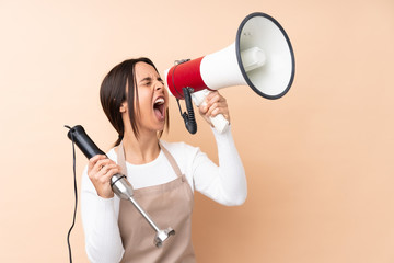 Young brunette girl using hand blender over isolated background shouting through a megaphone